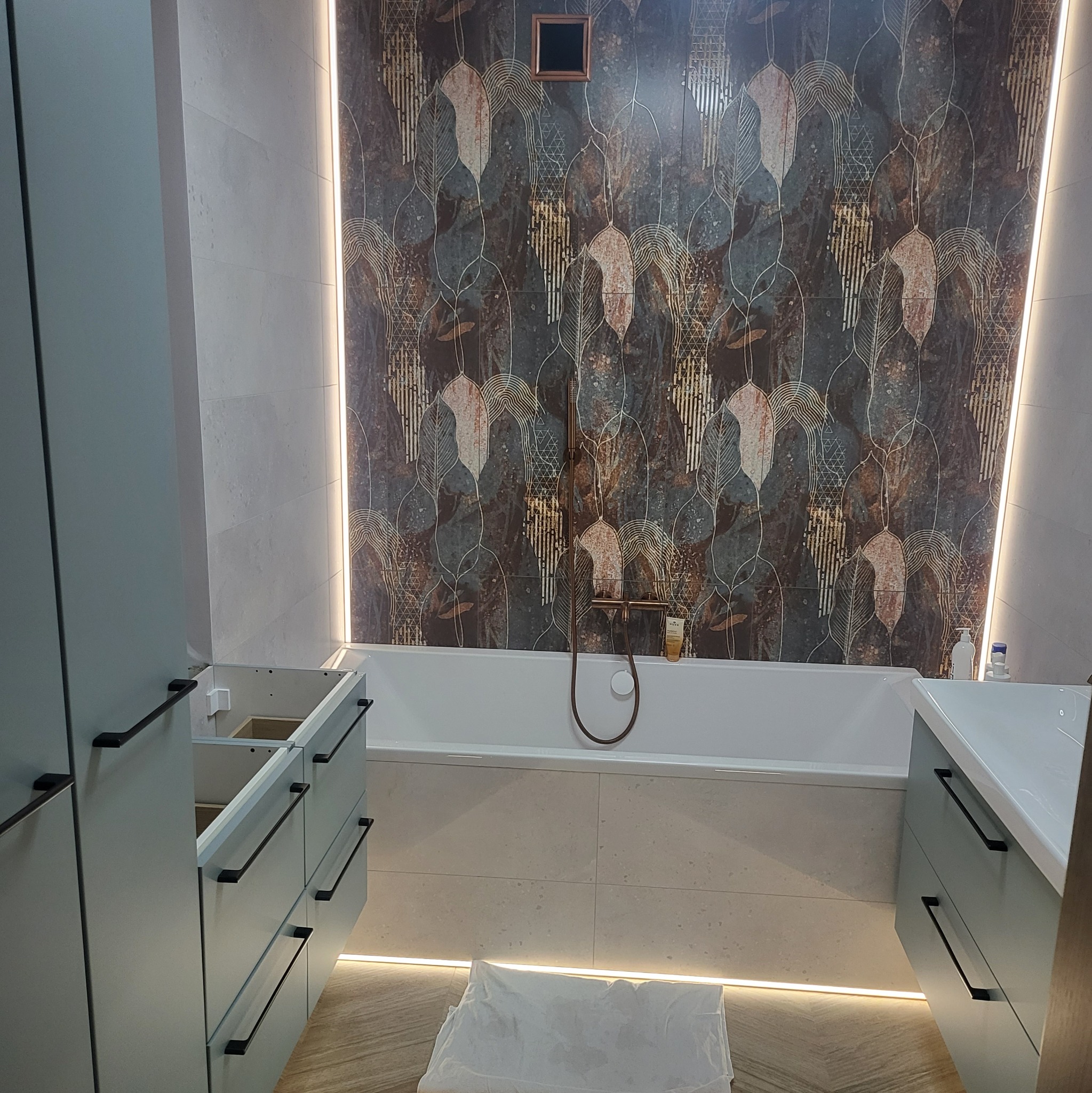 Nowoczesna łazienka w trakcie montażu: szafki pod umywalkę i wanna z baterią na tle wzorzystych płytek, oświetlenie LED pod szafkami i pionowe po bokach ściany.