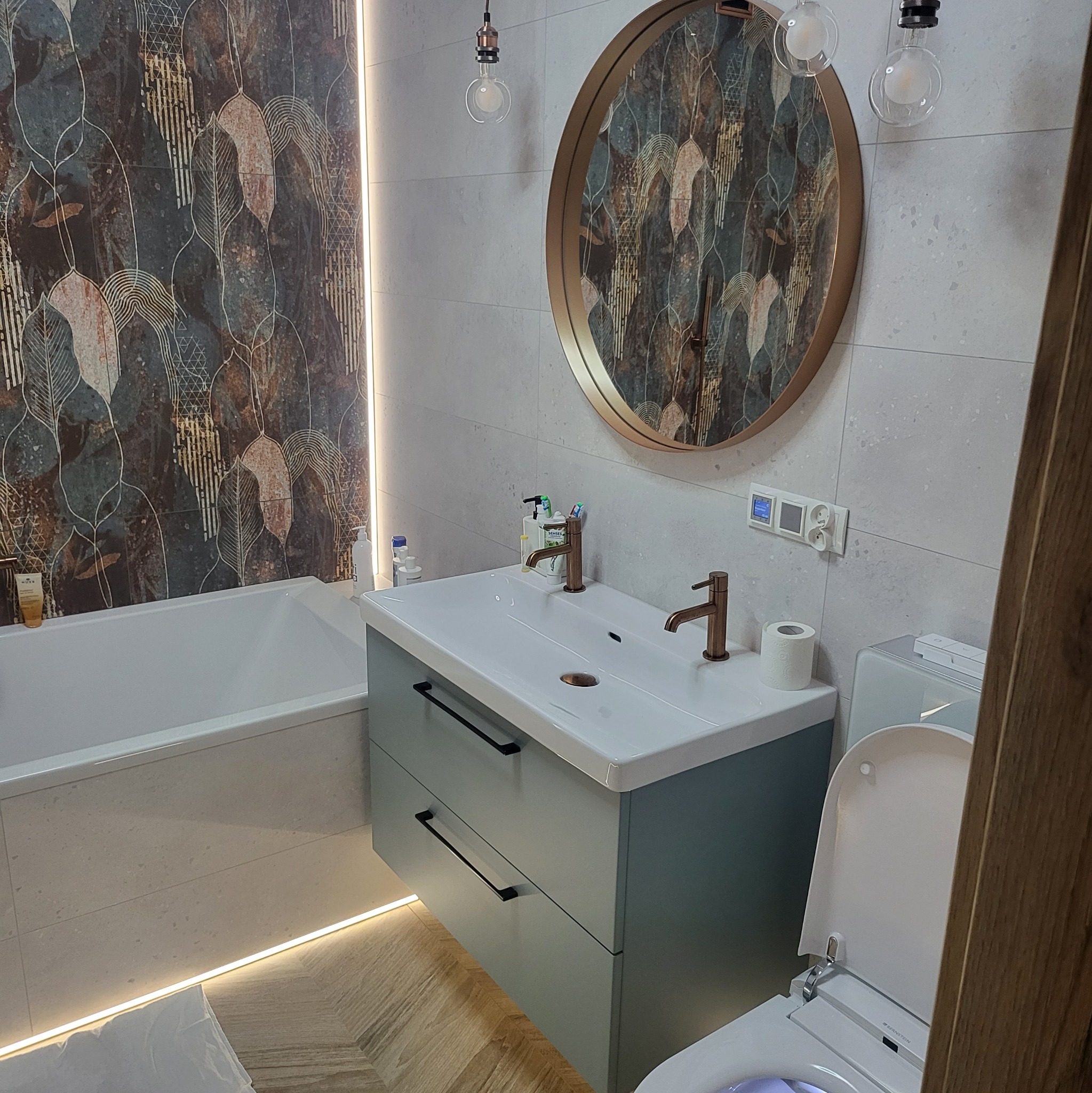 Nowoczesna łazienka z wanną, umywalką z szafką w kolorze gołębim, okrągłym lustrem w złotej ramie i dekoracyjnymi płytkami z motywem liści, oświetlona paskami LED.