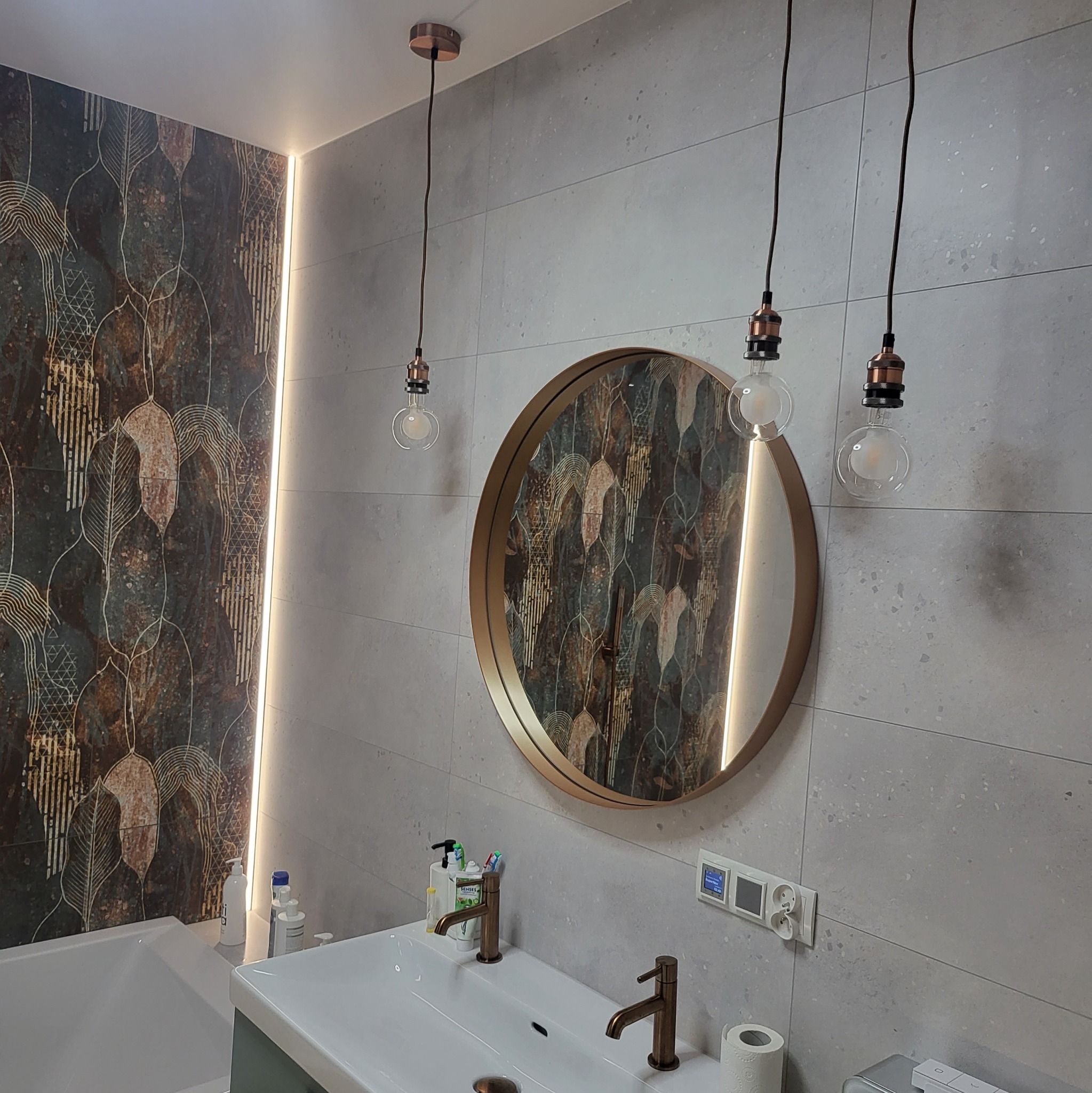 Nowoczesna łazienka z umywalką z dwoma kranami w kolorze miedzi, okrągłym lustrem w złotej ramie, trzema wiszącymi lampami z żarówkami Edisona i płytkami z motywem liści.