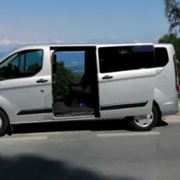 Srebrny van z otwartymi drzwiami przesuwnymi, zaparkowany na poboczu z widokiem na zalesione wzgórza w tle.