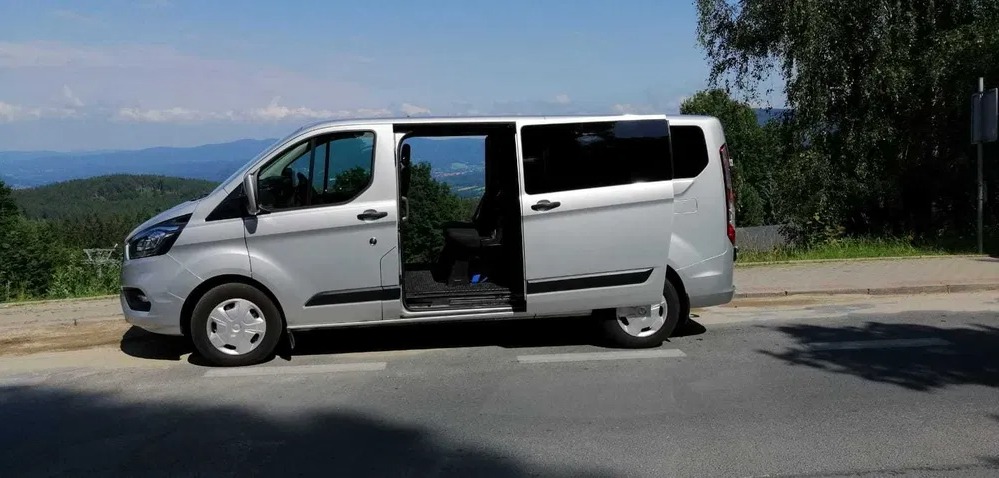 Srebrny van z otwartymi drzwiami przesuwnymi, zaparkowany na poboczu z widokiem na zalesione wzgórza w tle.