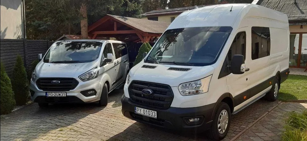 Dwa busy marki Ford, srebrny i biały, zaparkowane na brukowanym podjeździe przed domem z drewnianą wiatą.