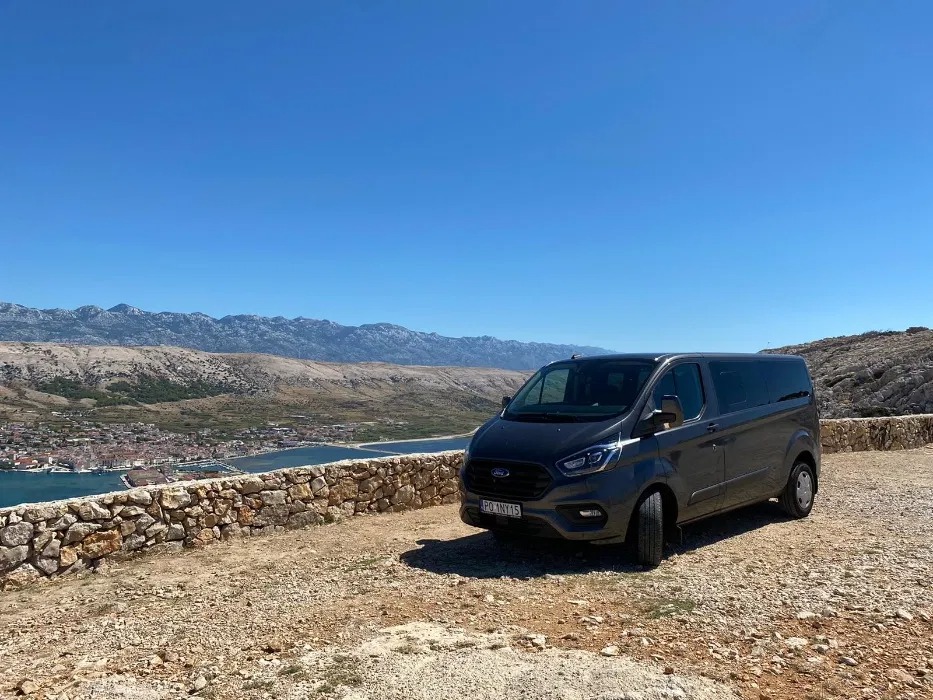 Szary van Ford Transit Custom na tle kamiennego murku, z widokiem na miasto, morze i góry w oddali, w słoneczny dzień.