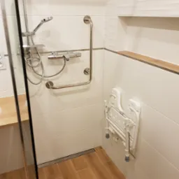Wykończona łazienka z prysznicem bez brodzika, drewnopodobnymi płytkami podłogowymi, białymi płytkami ściennymi z delikatną fakturą, uchwytami dla niepełnosprawnych i składanym siedziskiem...