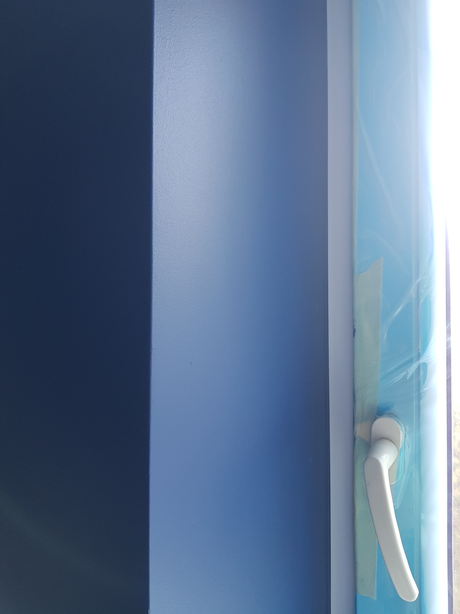 Świeżo pomalowana na niebiesko ściana z zamontowanym nowym oknem PCV, zabezpieczonym folią ochronną i taśmą malarską.