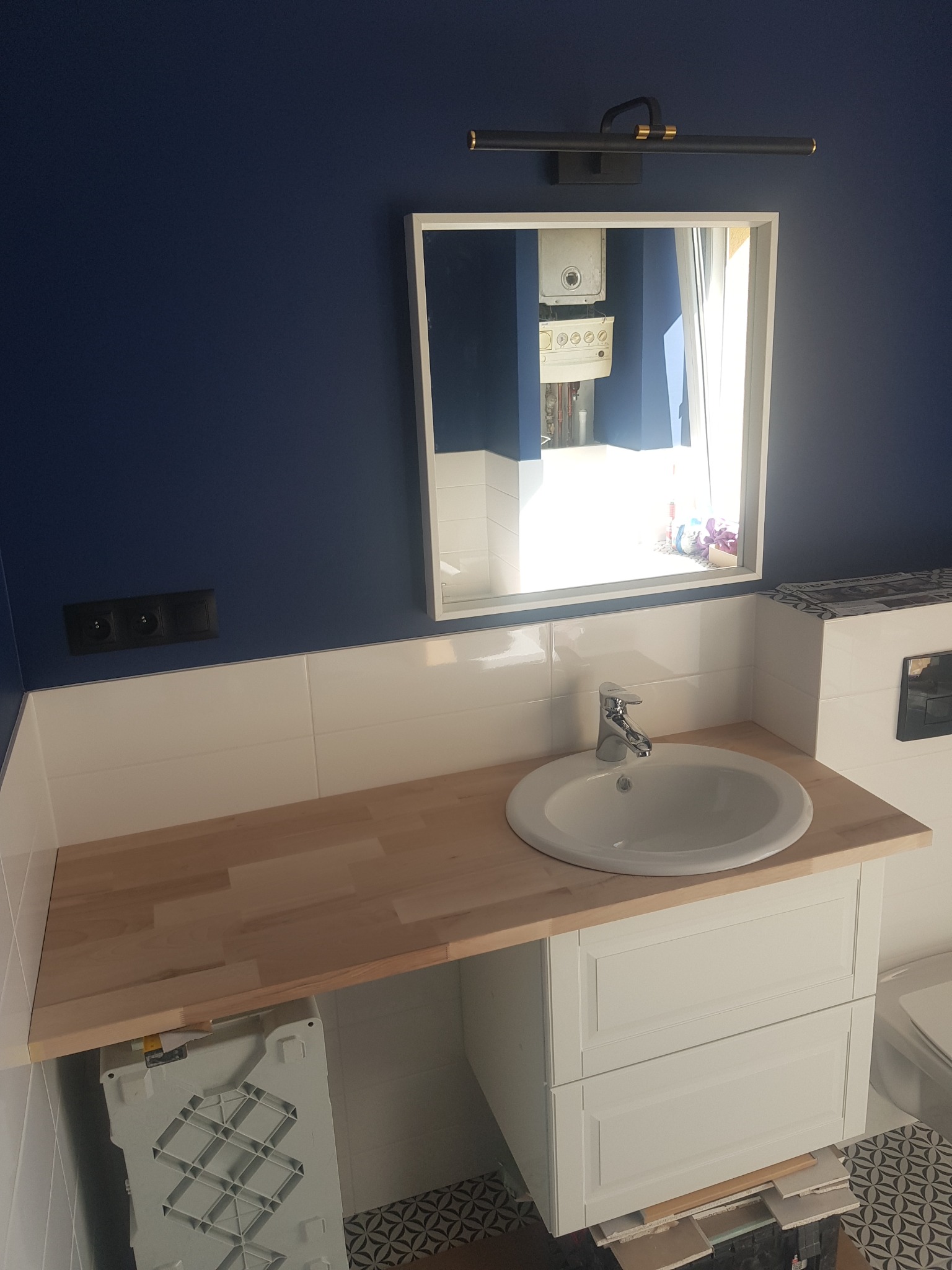 Umywalka z szafką i drewnianym blatem w łazience z granatową ścianą, widoczne białe płytki i lustro, w którym odbija się piec gazowy.