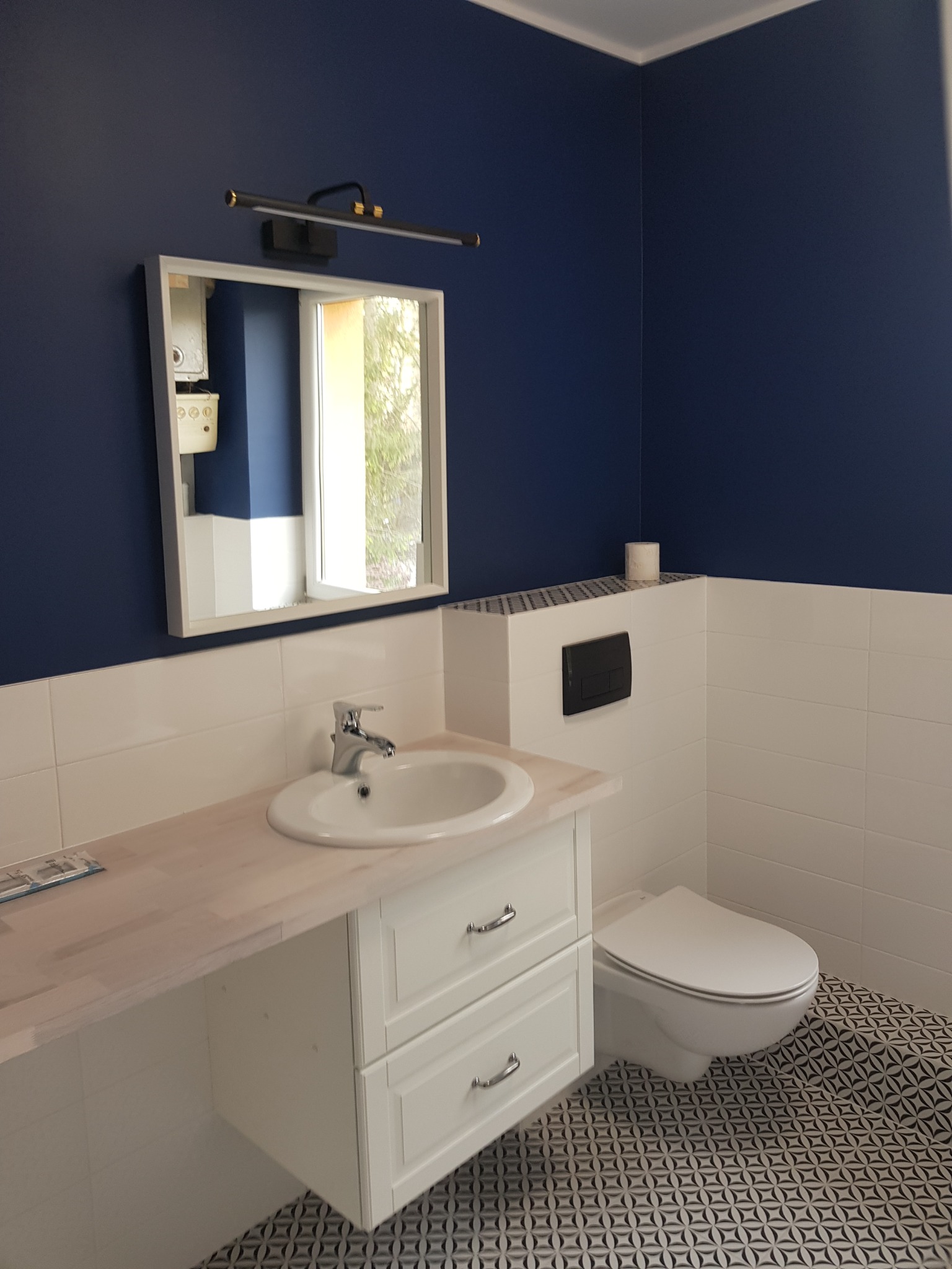 Nowoczesna łazienka z granatowymi ścianami, białymi płytkami, umywalką z szafką i geometryczną podłogą. W lustrze odbija się piec gazowy.