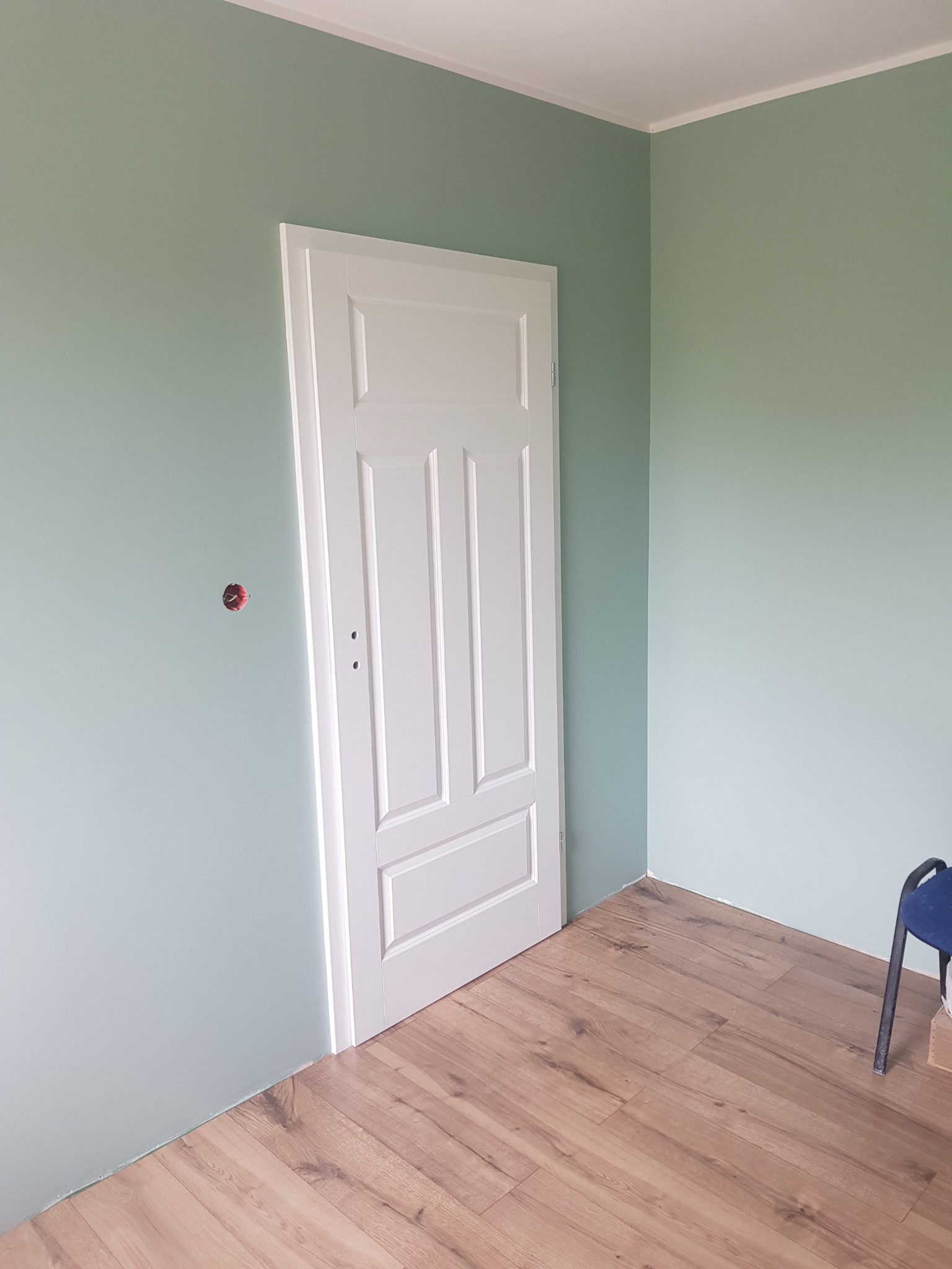 Świeżo pomalowane na zielono ściany w Szczecinie z nową, białą, trzydrzwiową ościeżnicą i panelami podłogowymi, widoczne gniazdko elektryczne bez osłony.