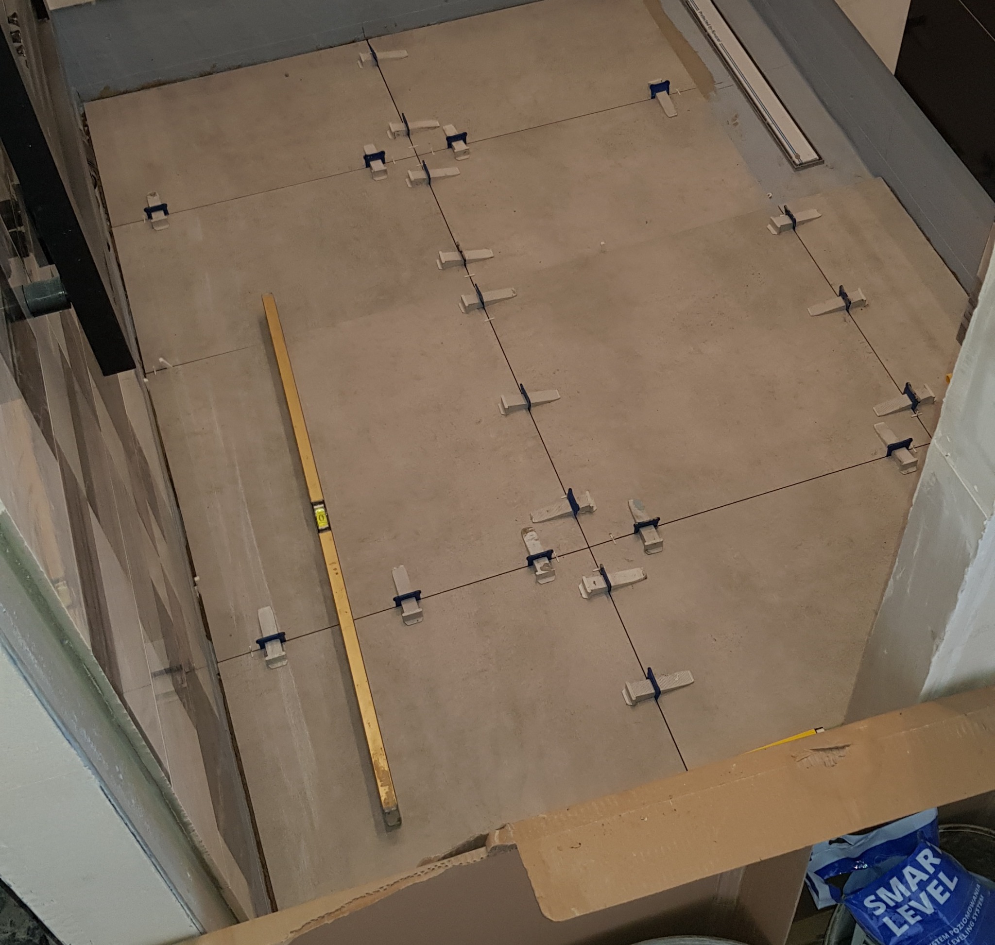 Układanie beżowych płytek podłogowych z użyciem systemu poziomowania, widoczny poziomica przy ścianie i opakowanie po systemie poziomowania.