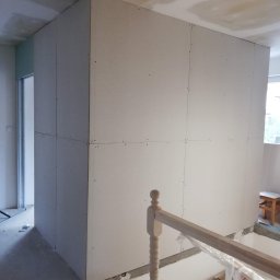 RAFEXAN RAFAŁ HELIASZ - Wnętrze w trakcie prac: montaż płyt gipsowo-kartonowych na ścianach, widoczne profile i drewniana balustrada schodów w surowym stanie.