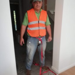 RAFEXAN RAFAŁ HELIASZ - Mężczyzna w kamizelce odblaskowej stoi w przejściu między pokojami, z widoczną nową podłogą w jednym pomieszczeniu i przygotowanym do remontu pokojem w tle.