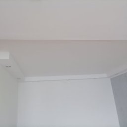 RAFEXAN RAFAŁ HELIASZ - Wnętrze po remoncie: białe ściany, sufit podwieszany z oświetleniem punktowym, fragment ściany w odcieniach szarości.