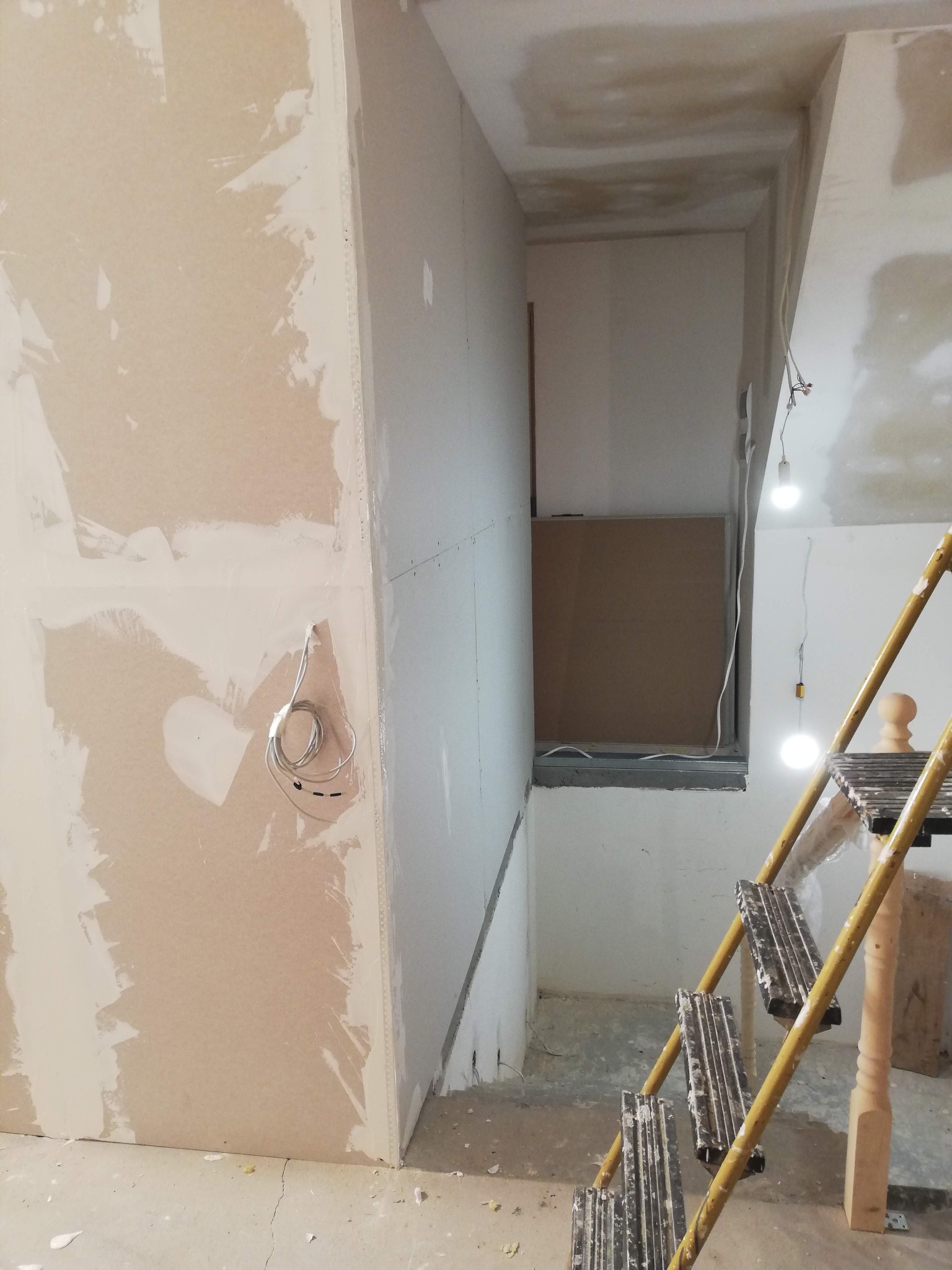 Wnętrze w trakcie prac wykończeniowych: ściany z płyt gipsowo-kartonowych z widocznymi łączeniami, żółta drabina malarska i prowizoryczne oświetlenie z żarówek na kablach.