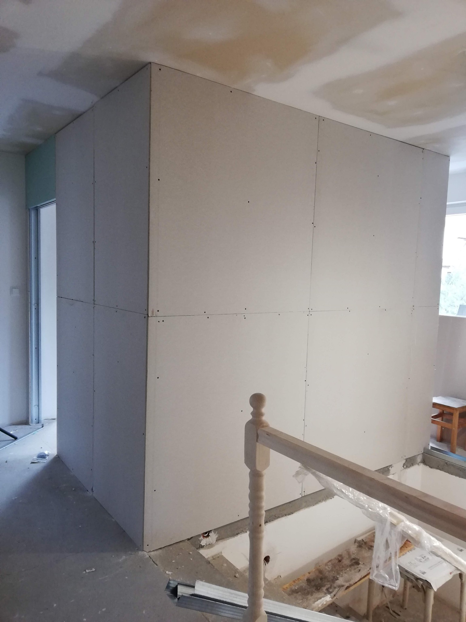 Wnętrze w trakcie prac: montaż płyt gipsowo-kartonowych na ścianach, widoczne profile i drewniana balustrada schodów w surowym stanie.