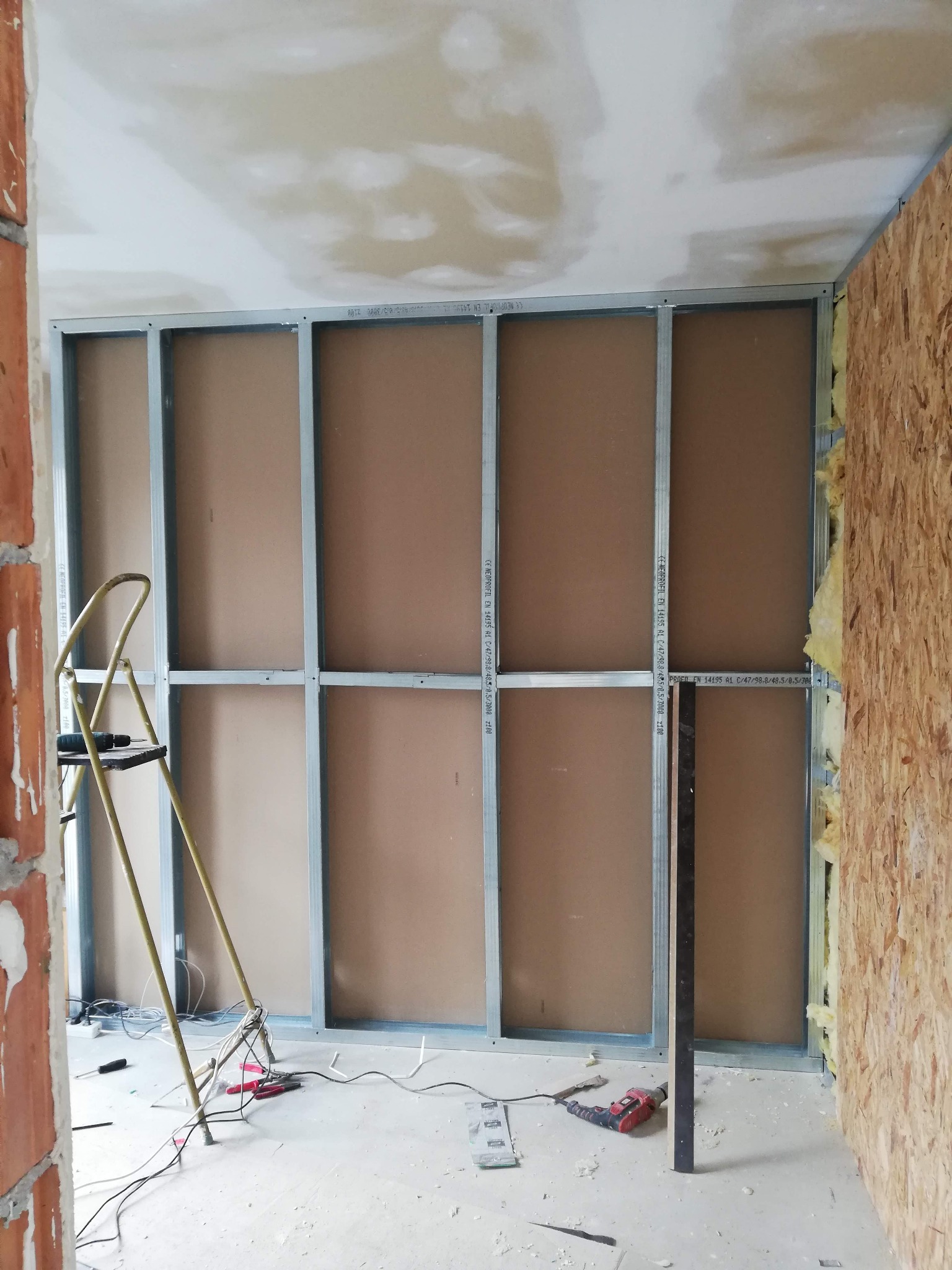 Wnętrze w trakcie prac wykończeniowych: metalowy stelaż pod płyty gipsowo-kartonowe, częściowo obłożony płytami, widoczna drabina, narzędzia i kable na podłodze, ściana z płyty OSB z widoczną...