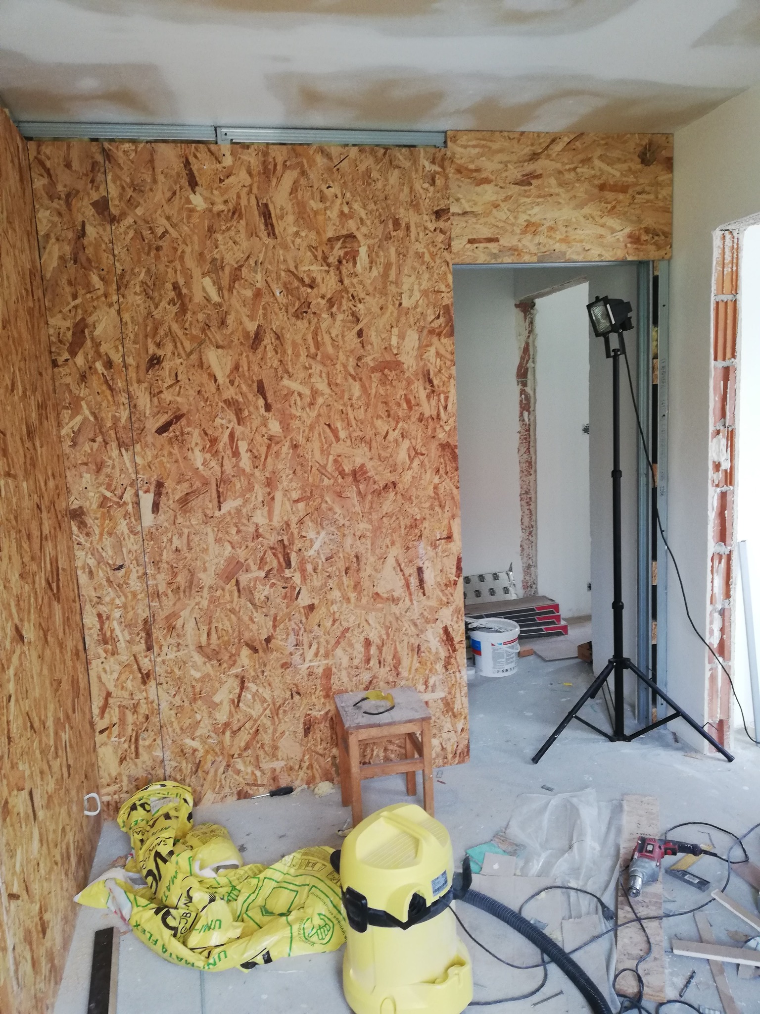 Wnętrze w trakcie prac remontowych: ściana wyłożona płytą OSB, metalowy profil sufitowy, lampa na statywie, odkurzacz przemysłowy, narzędzia i materiały budowlane na podłodze.