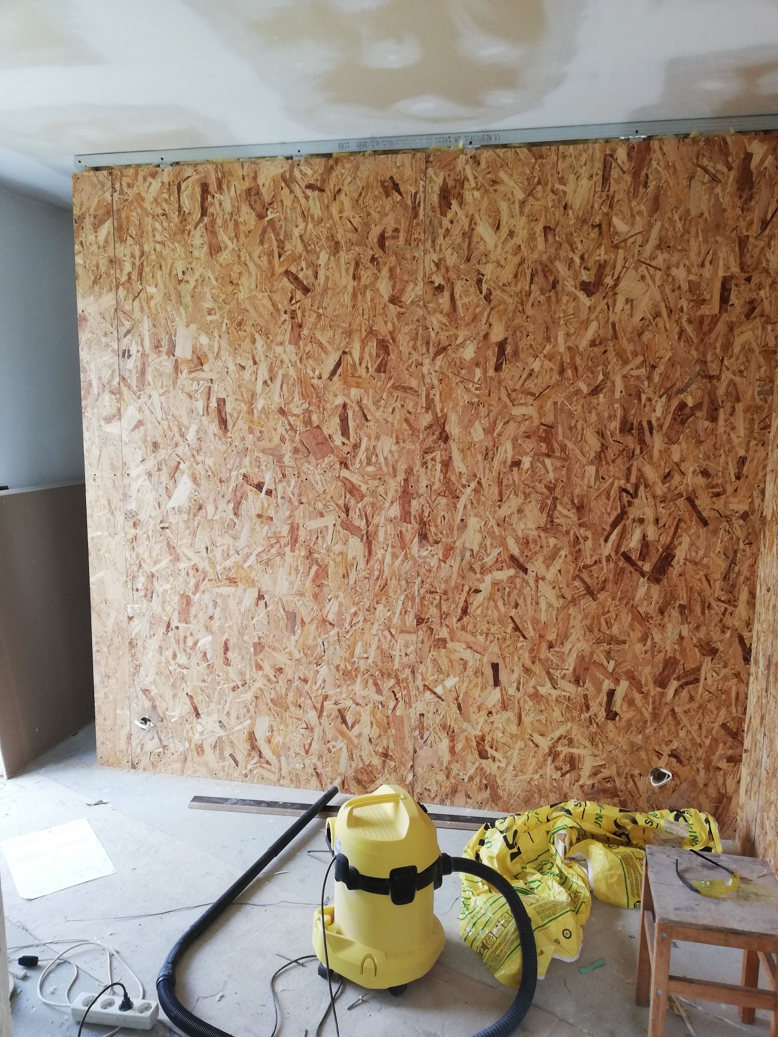 Ściana z płyty OSB w trakcie montażu, widoczne metalowe profile mocujące, żółty odkurzacz przemysłowy i materiały izolacyjne na podłodze.
