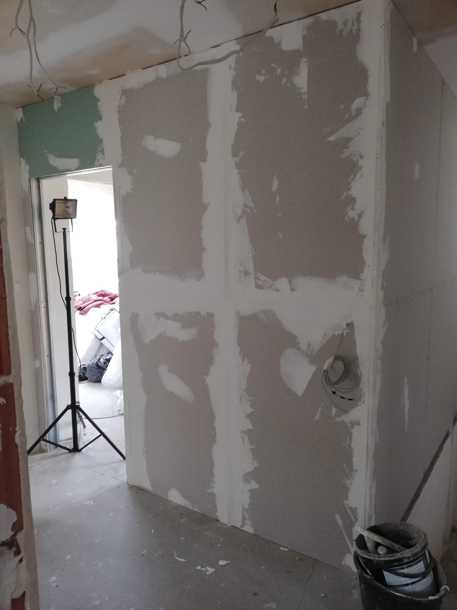 Ściana z płyt gipsowo-kartonowych w trakcie szpachlowania, widoczne łączenia płyt i instalacja elektryczna, oświetlenie budowlane na trójnogu w tle, wiadro z narzędziami przy ścianie.