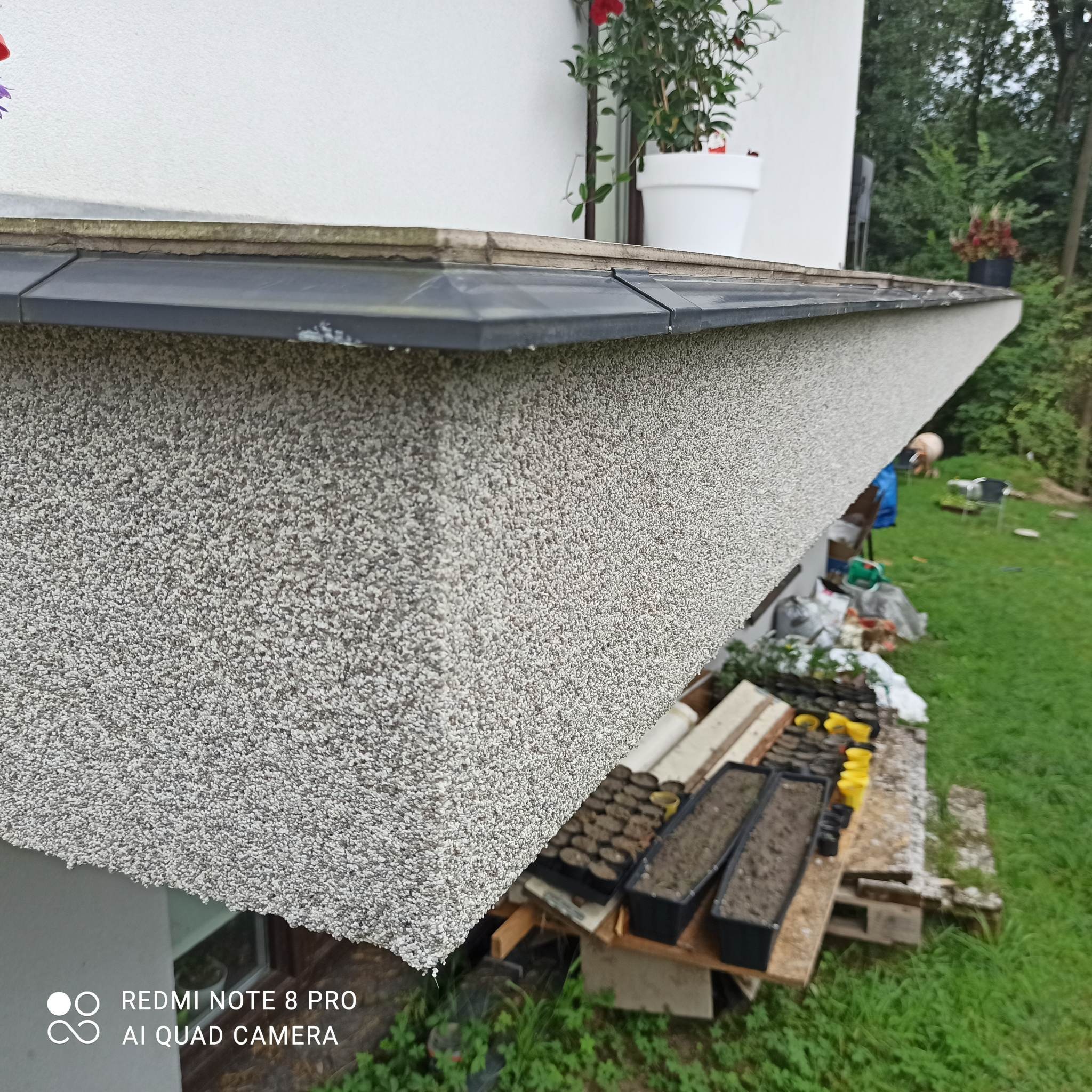 Fragment elewacji budynku z fakturą baranka, pod nią paleta z sadzonkami i narzędziami ogrodniczymi, w tle trawnik i zieleń.