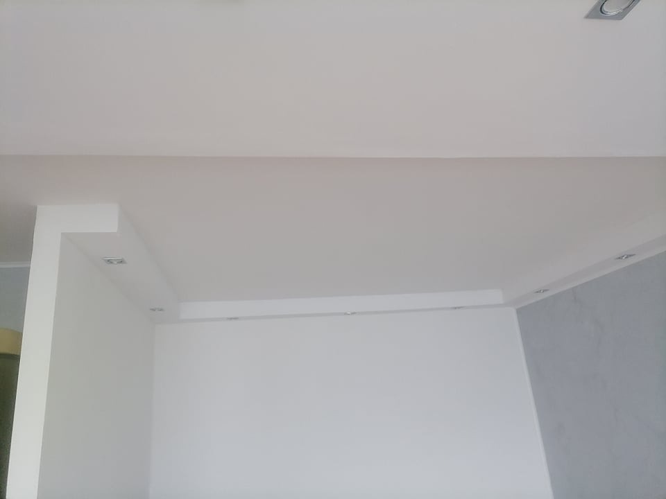 Wnętrze po remoncie: białe ściany, sufit podwieszany z oświetleniem punktowym, fragment ściany w odcieniach szarości.