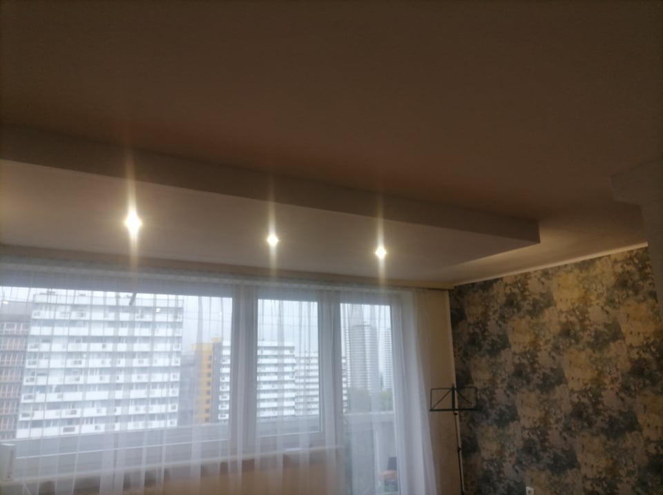 Pokój z oknem, przez które widać wysokie budynki, ściana pokryta tapetą w odcieniach szarości i beżu, podwieszany sufit z trzema punktowymi lampami.