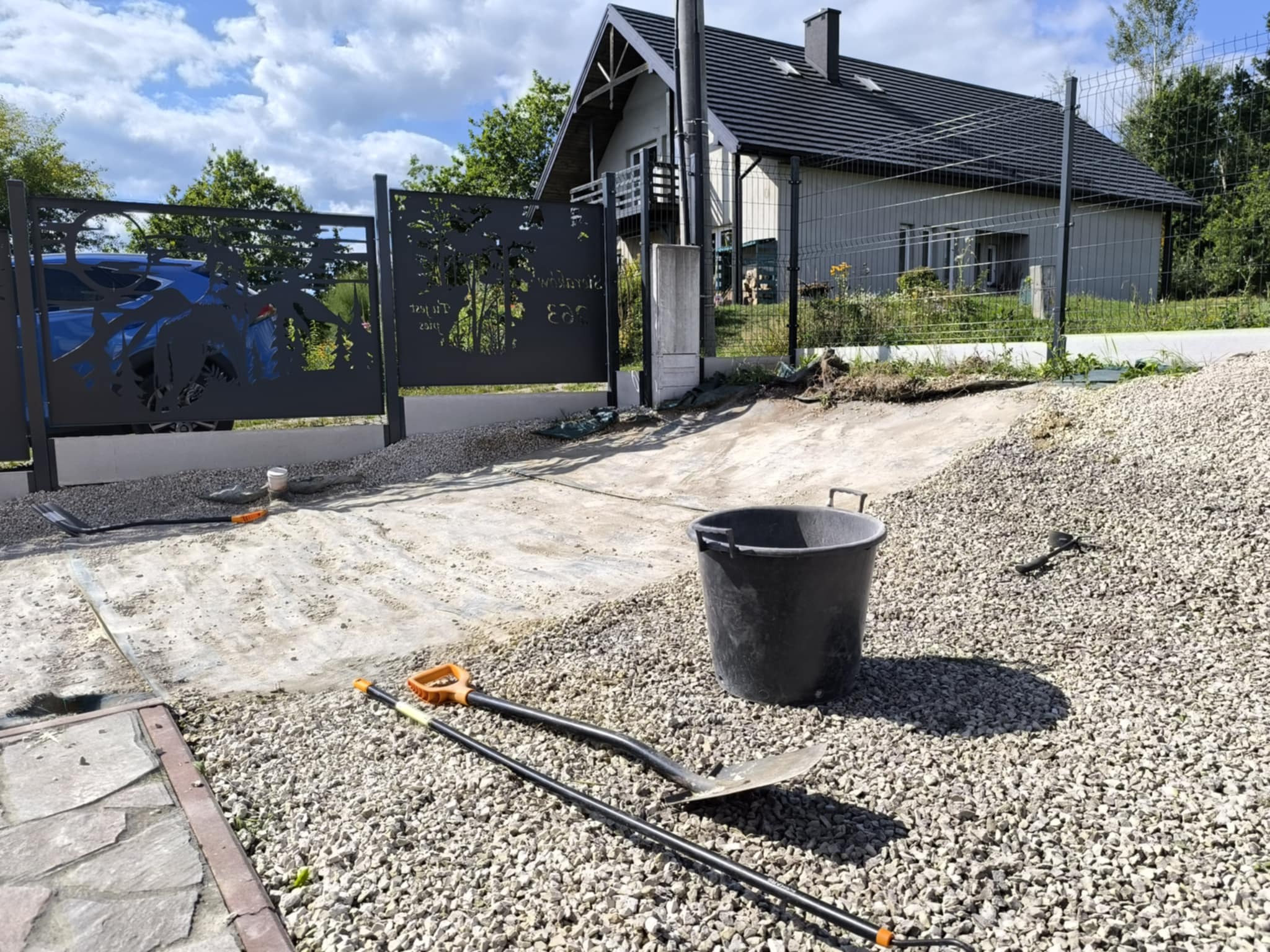 Prace przygotowawcze do ułożenia nawierzchni z kamienia przy posesji z dekoracyjnym ogrodzeniem. Widoczne narzędzia i wiadro.