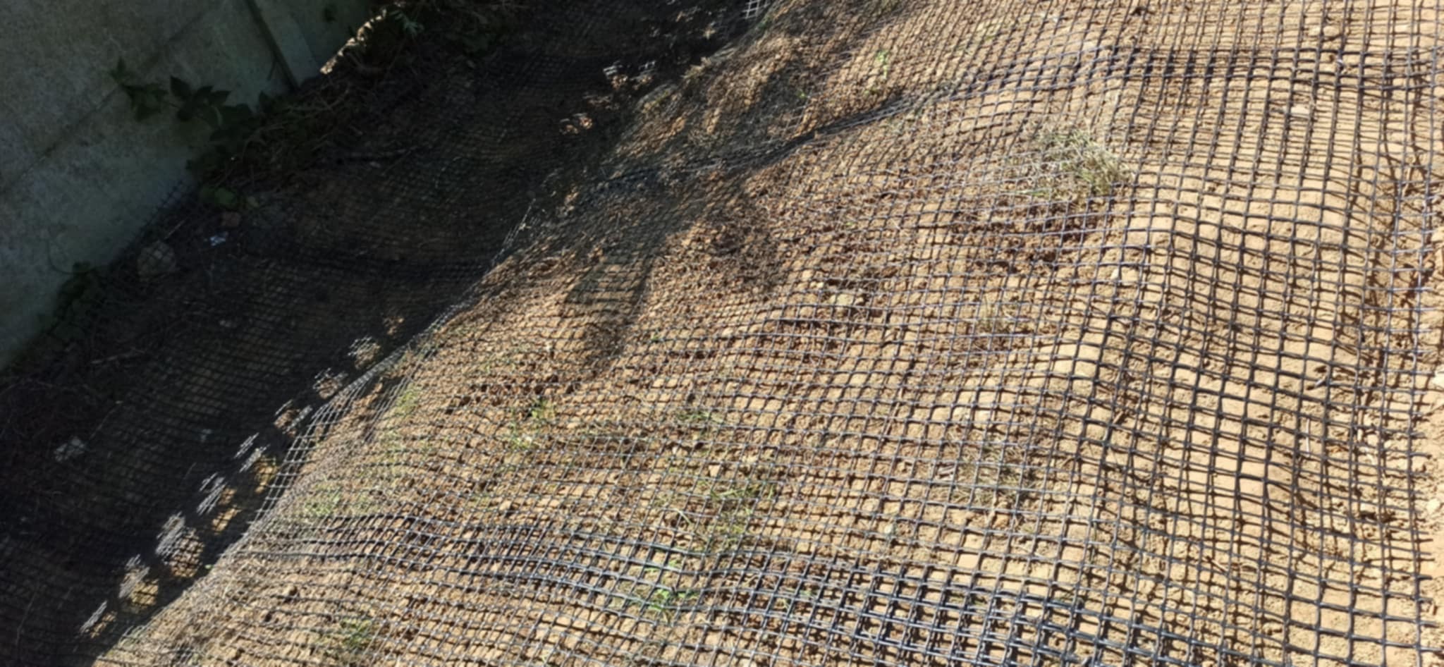 Siatka wzmacniająca skarpę z piasku, widok z góry pod kątem. Element stabilizacji gruntu na zboczu, przygotowany do dalszych prac ogrodniczych.