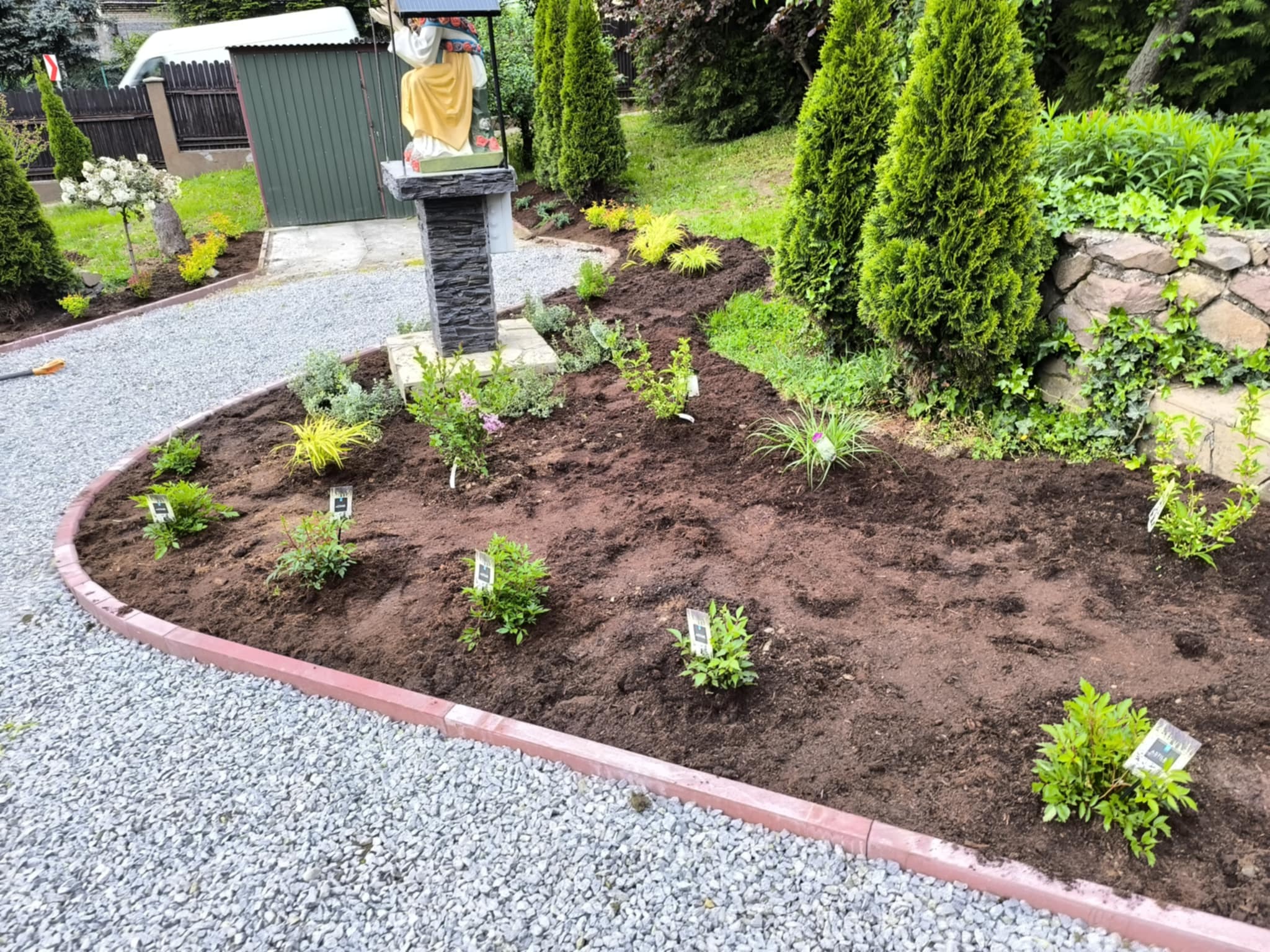 Świeżo posadzone, młode rośliny w ogrodzie z brązową ziemią, obok żwirowa ścieżka z czerwoną obwódką i dekoracyjna statuetka.