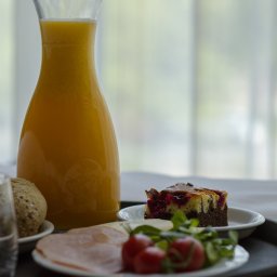 FHU ''CERTEX'' Jadwiga Świętalska - Śniadanie na tacy: dzbanek soku pomarańczowego, bułka, plasterki szynki, pomidorki koktajlowe i kawałek ciasta z wiśniami na białych talerzykach.