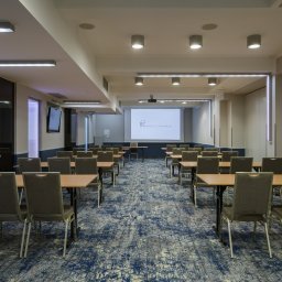 FHU ''CERTEX'' Jadwiga Świętalska - Sala konferencyjna z rzędami stołów i krzeseł, projektor wyświetla logo hotelu na ekranie, niebieski wzorzysty dywan na podłodze.