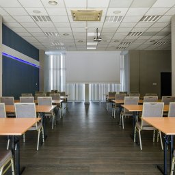 FHU ''CERTEX'' Jadwiga Świętalska - Sala konferencyjna z rzędami stołów i krzeseł, projektor zamontowany na suficie, roleta ekranu opuszczona, oświetlenie LED wzdłuż ściany.