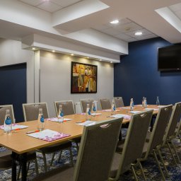 FHU ''CERTEX'' Jadwiga Świętalska - Sala konferencyjna z długim stołem, krzesłami, butelkami wody i notatnikami, ściana w kolorze granatowym z telewizorem, obraz na ścianie w tle.