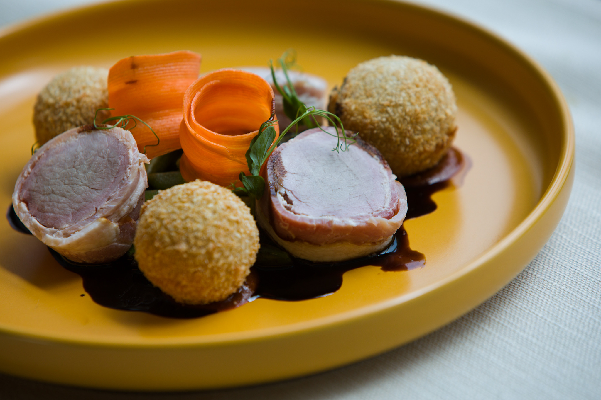 Elegancko podane danie: medaliony wieprzowe owinięte boczkiem, krokiety i dekoracja z marchewki na żółtym talerzu z ciemnym sosem.