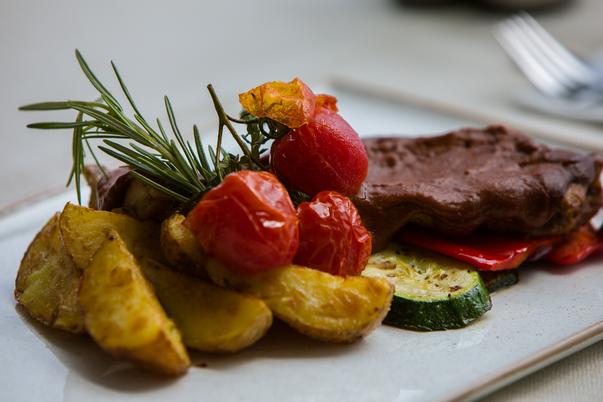 Elegancko podane danie: pieczone ziemniaki, grillowana cukinia, pomidorki cherry i mięso z sosem, udekorowane gałązką rozmarynu.