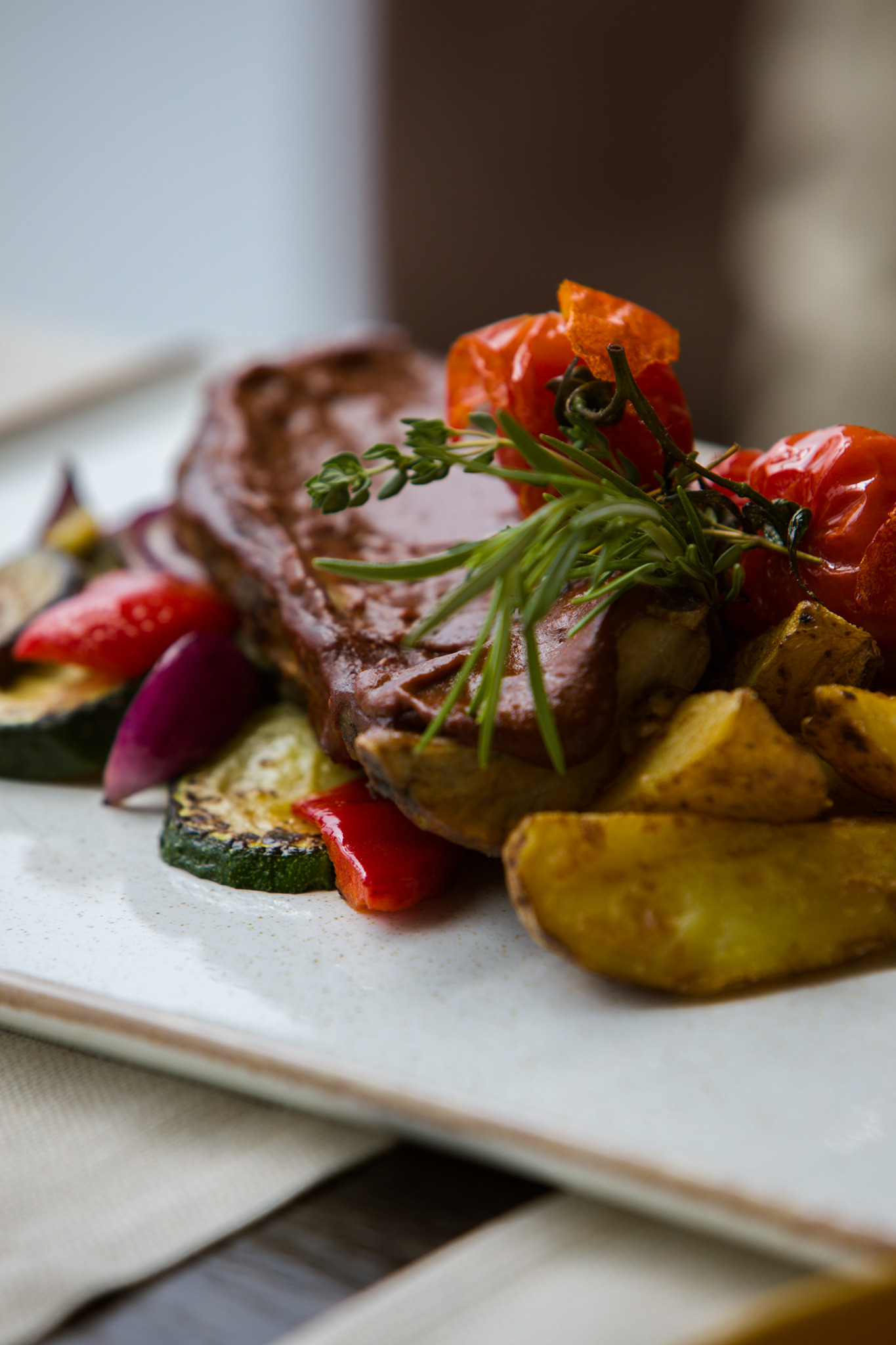 Elegancko podane danie: stek w ciemnym sosie, otoczony grillowanymi warzywami (cukinia, papryka, czerwona cebula), pieczonymi ziemniakami i pomidorkami cherry, udekorowane gałązką rozmarynu...