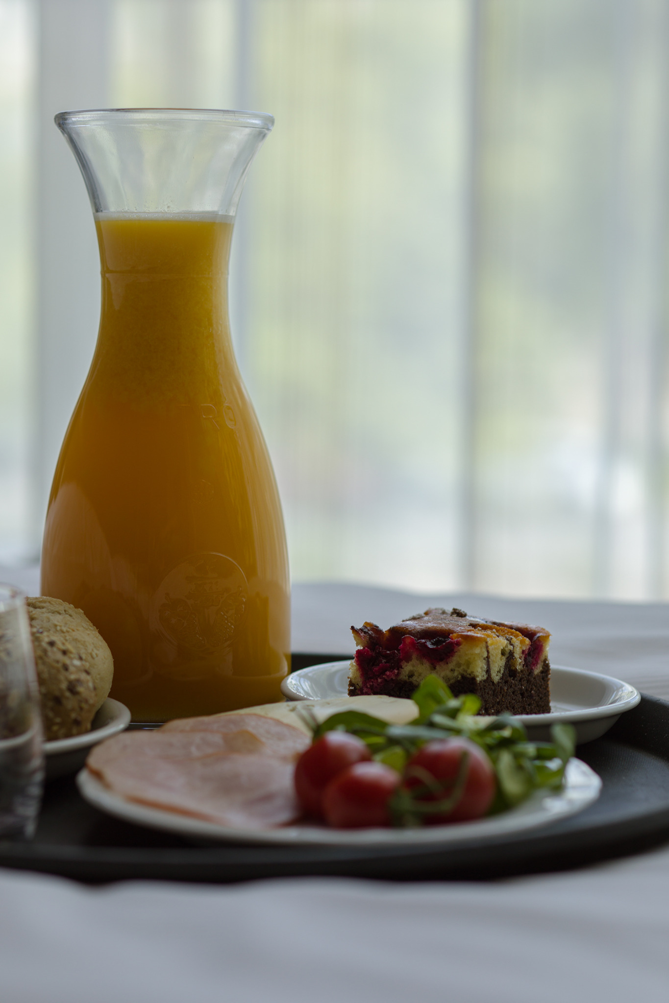 Śniadanie na tacy: dzbanek soku pomarańczowego, bułka, plasterki szynki, pomidorki koktajlowe i kawałek ciasta z wiśniami na białych talerzykach.