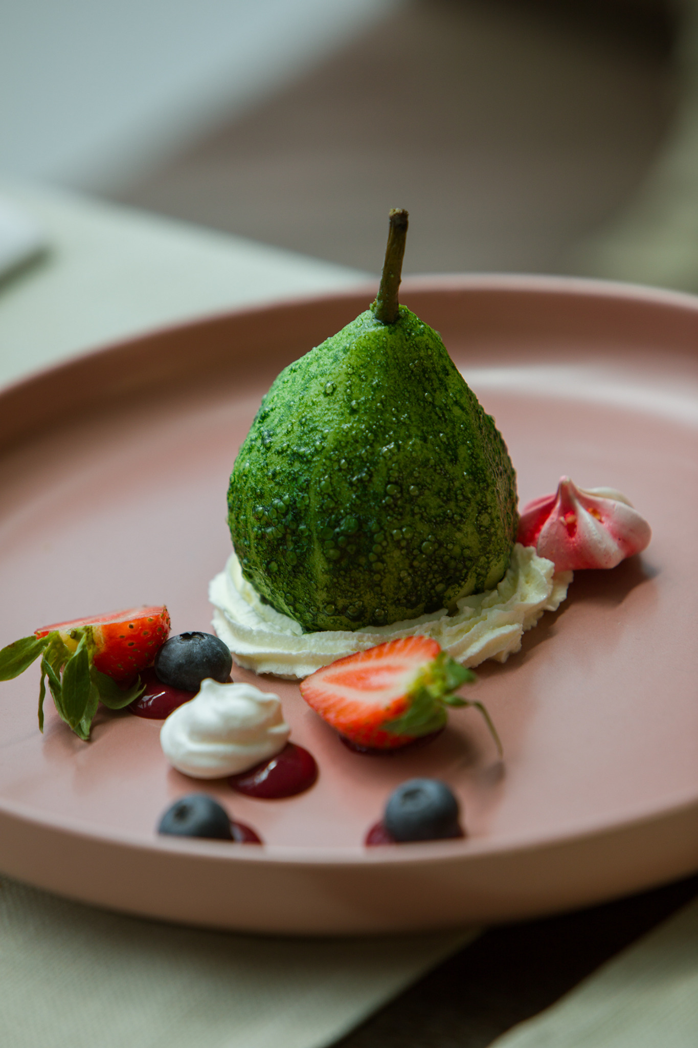 Deser: zielona gruszka obłożona bitą śmietaną, udekorowana truskawkami, borówkami i bezami na różowym talerzu.