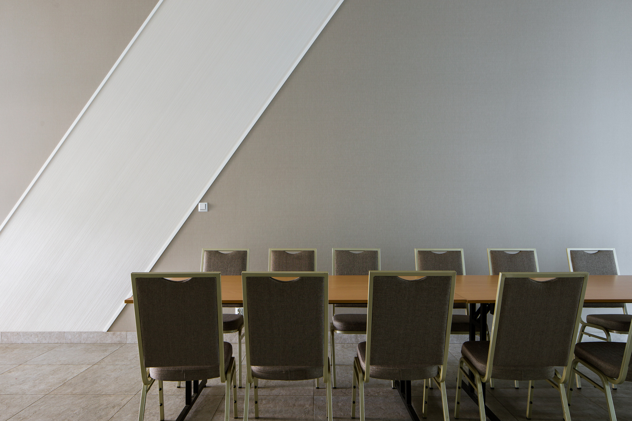 Sala konferencyjna z długim drewnianym stołem i ustawionymi wokół niego krzesłami z szarą tapicerką, na ścianie widoczny element dekoracyjny w postaci białego trójkąta.