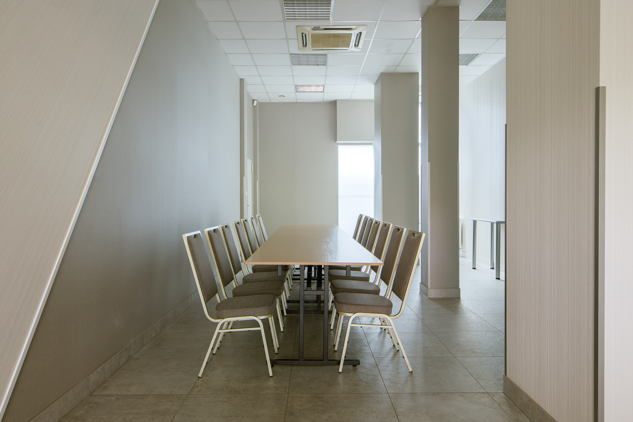 Sala konferencyjna z długim stołem i krzesłami w odcieniach szarości i beżu, widok z perspektywy wejścia, sufit kasetonowy z klimatyzacją.