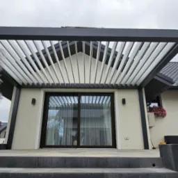 Nowoczesna, czarno-biała konstrukcja z regulowanymi lamelami nad tarasem i przesuwnymi drzwiami, z widoczną roślinnością ozdobną i elementami wykończenia elewacji.