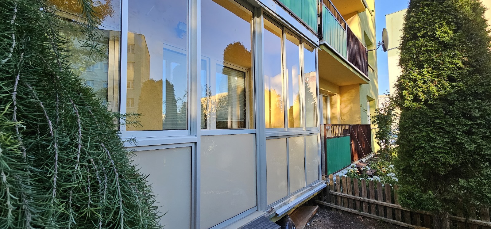 Zabudowany balkon z aluminiowymi ramami i częściowo matowymi panelami, widok z zewnątrz, z krzewem rozmarynu na pierwszym planie i odbiciem budynku w szybach.