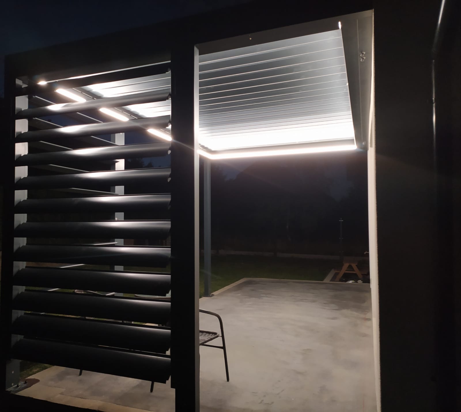 Nowoczesna konstrukcja z metalowymi lamelami i oświetleniem LED nocą, widok na betonowy taras i ogród.