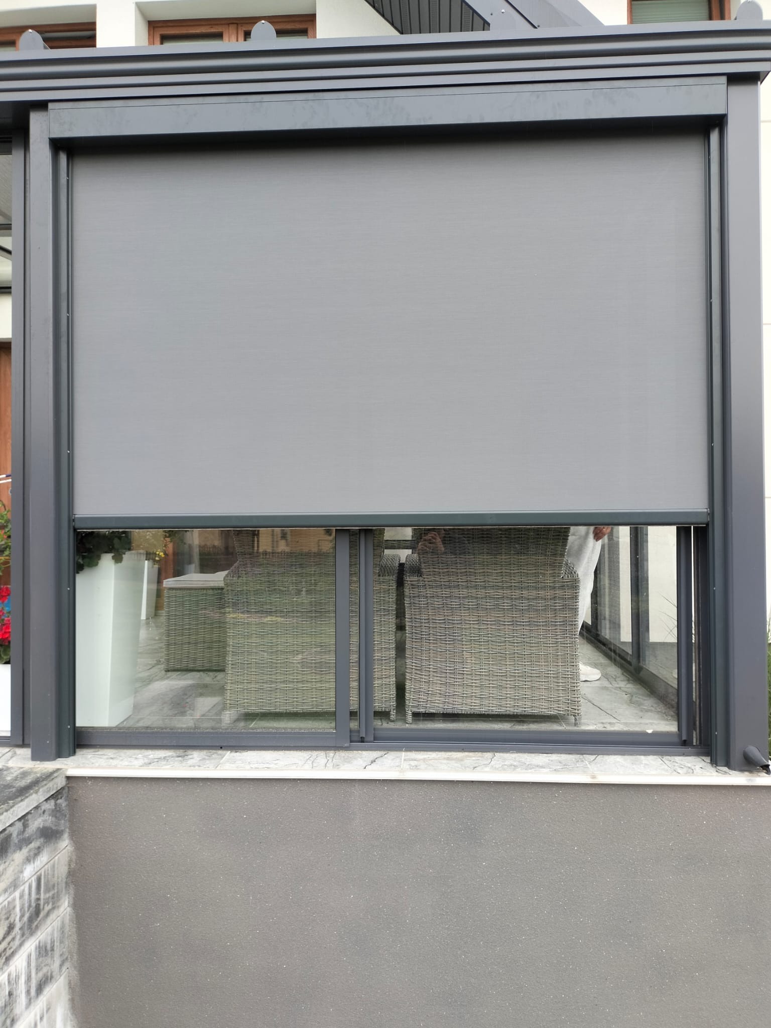 Zasłonięta roleta w metalowej pergoli, widoczne meble rattanowe na zewnątrz, szara konstrukcja i ściana.
