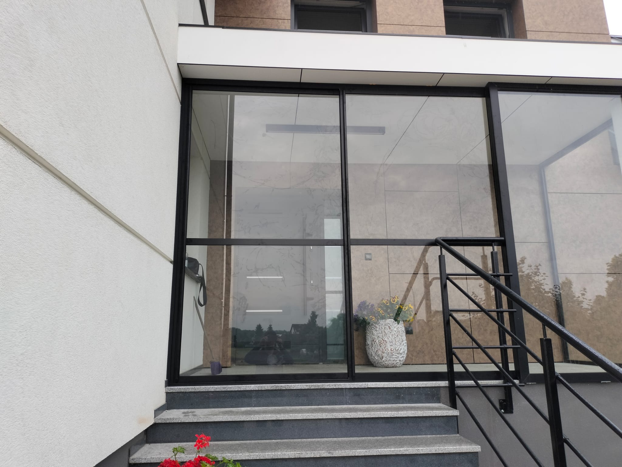 Nowoczesne wejście do domu z przeszklonymi drzwiami przesuwnymi w czarnej ramie, widoczne schody z szarego kamienia oraz kwiaty w ozdobnej donicy.