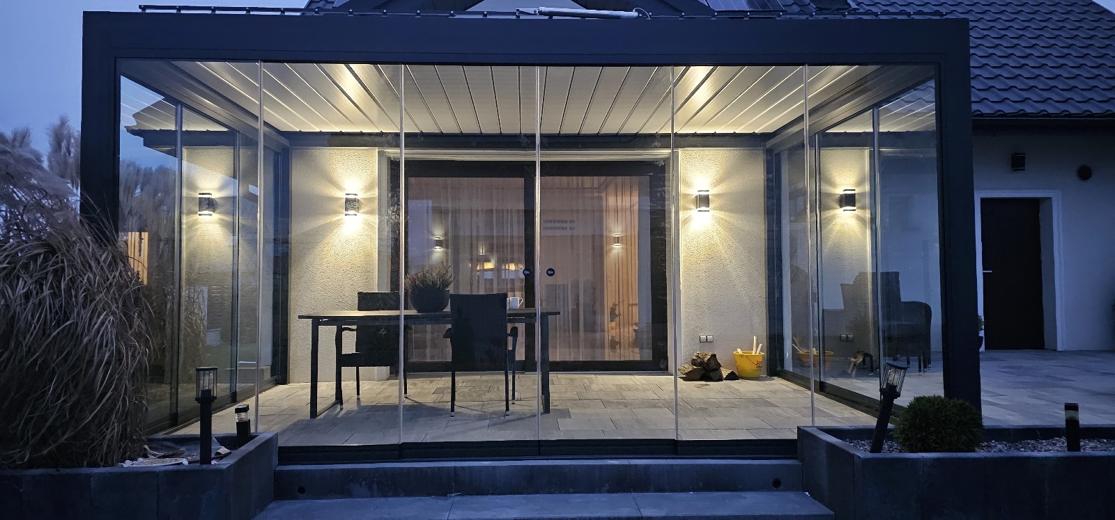Zadaszona przestrzeń tarasowa z metalową konstrukcją, osłonięta szklanymi panelami przesuwnymi, wyposażona w stół i krzesła, oświetlona zewnętrznymi lampami ściennymi, z widocznymi elementami...