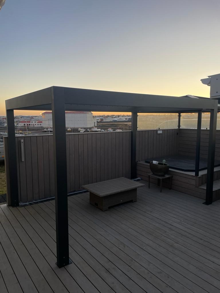 Nowoczesna metalowa pergola z przeszklonymi bokami, z jacuzzi na drewnianym tarasie i stolikiem kawowym, widok na miasto o zmierzchu.