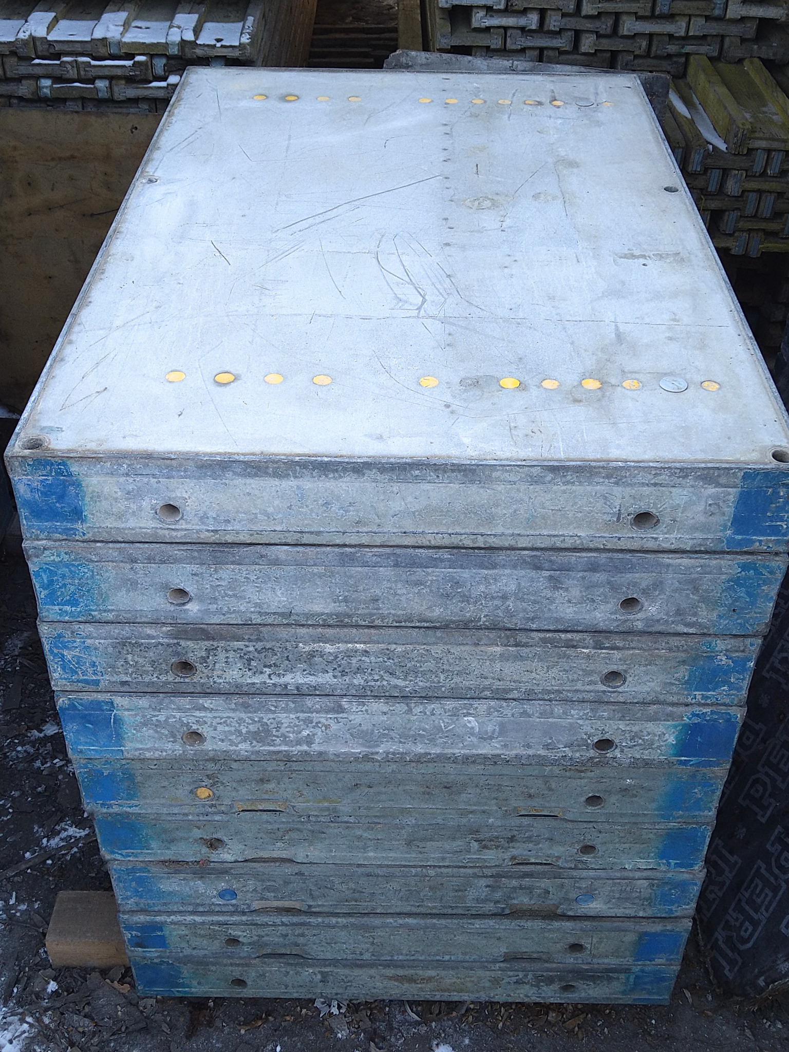 Stos szalunków budowlanych z widocznymi śladami użytkowania, niebieskimi krawędziami i żółtymi otworami, ułożony na ziemi z resztkami śniegu.