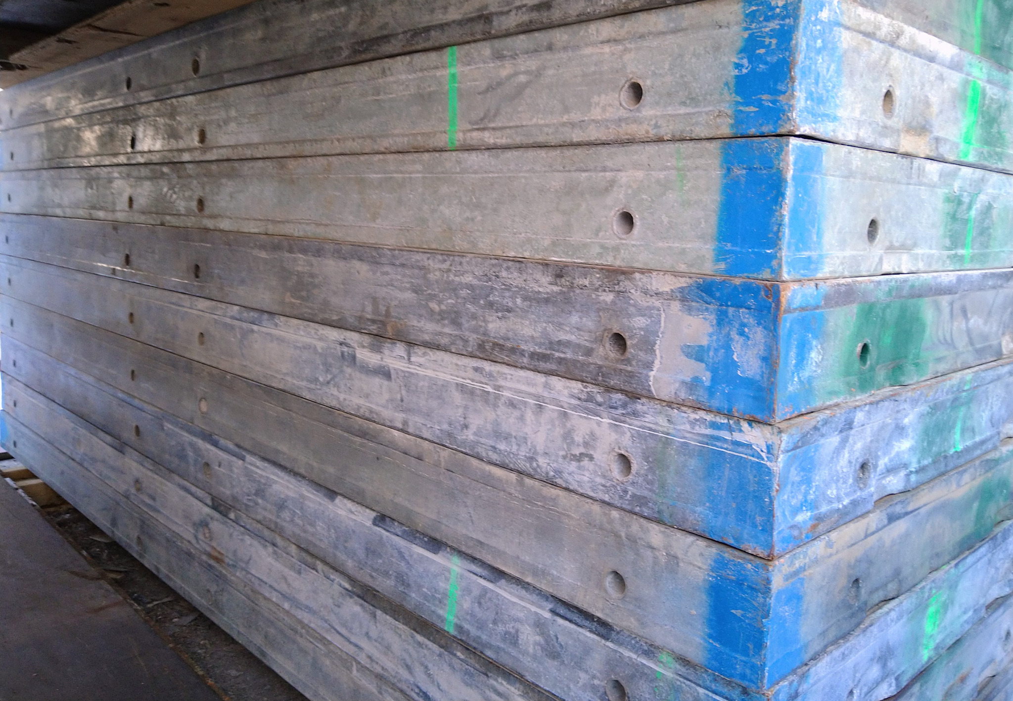 Stos szalunków budowlanych pomalowanych na niebiesko i szaro, z widocznymi otworami i zielonymi oznaczeniami, ułożonych na palecie.