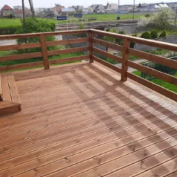 Nowy taras z desek ryflowanych, z drewnianą balustradą, widok na zielony ogr&oacute;d i stację kolejową w oddali.