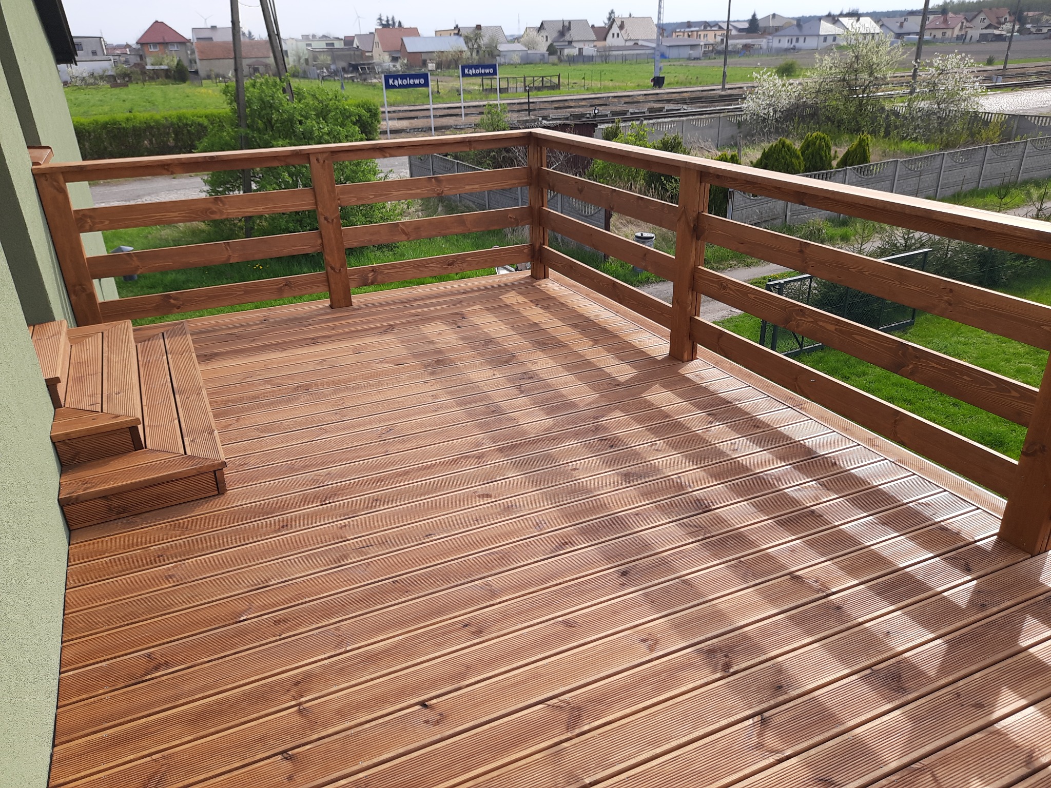 Nowy taras z desek ryflowanych, z drewnianą balustradą, widok na zielony ogród i stację kolejową w oddali.